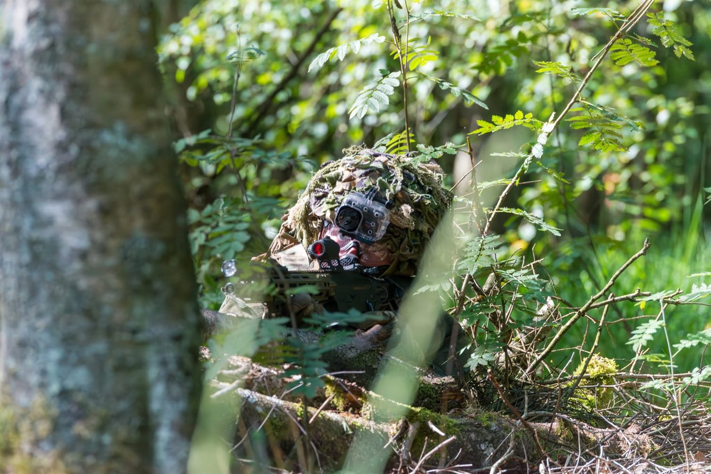 Camoufleret spiller med kamera på hjelmen ligger skjult bag grene og rønnebærløv