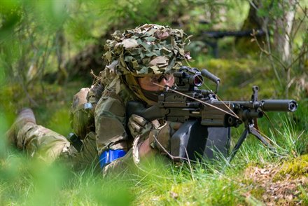Spiller med camouflagenet på hjelmen ligger med støttevåben på tofod i det høje græs
