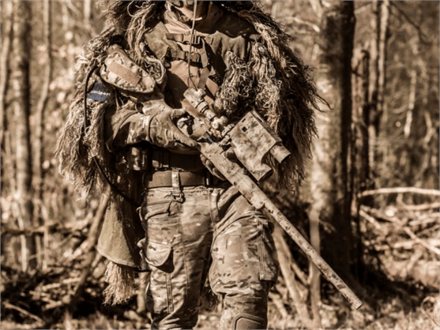 Spiller i ghillie-dragt med skarpskytteriffel mellem træerne